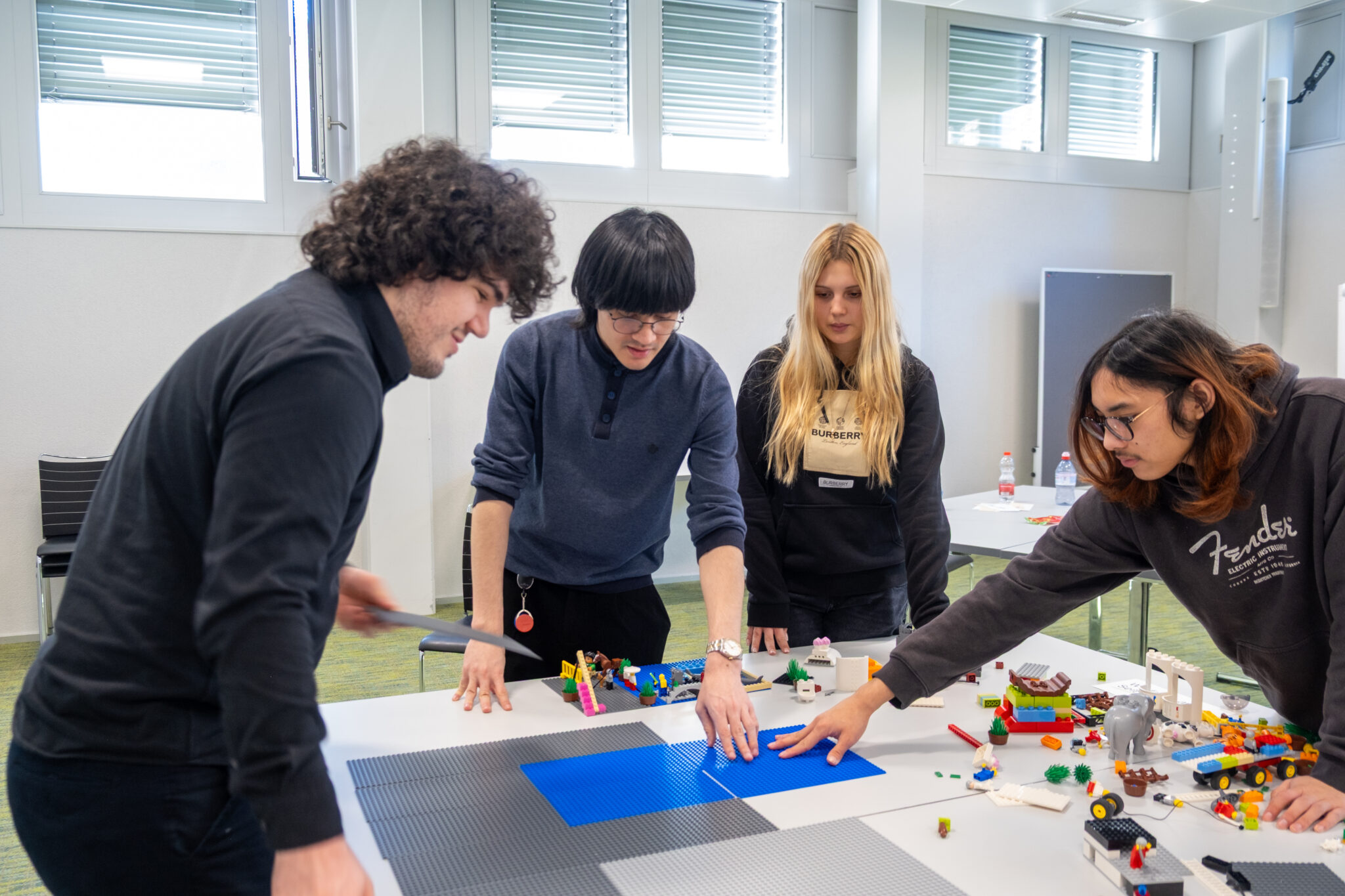 Vier junge Erwachsene arbeiten gemeinsam an einem Tisch und ordnen LEGO-Steine und -Figuren auf einem Raster aus blauen und grauen Matten in einem hellen Klassenzimmer an. Sie wirken konzentriert und sind ganz in eine praktische Gruppenaktivität vertieft.