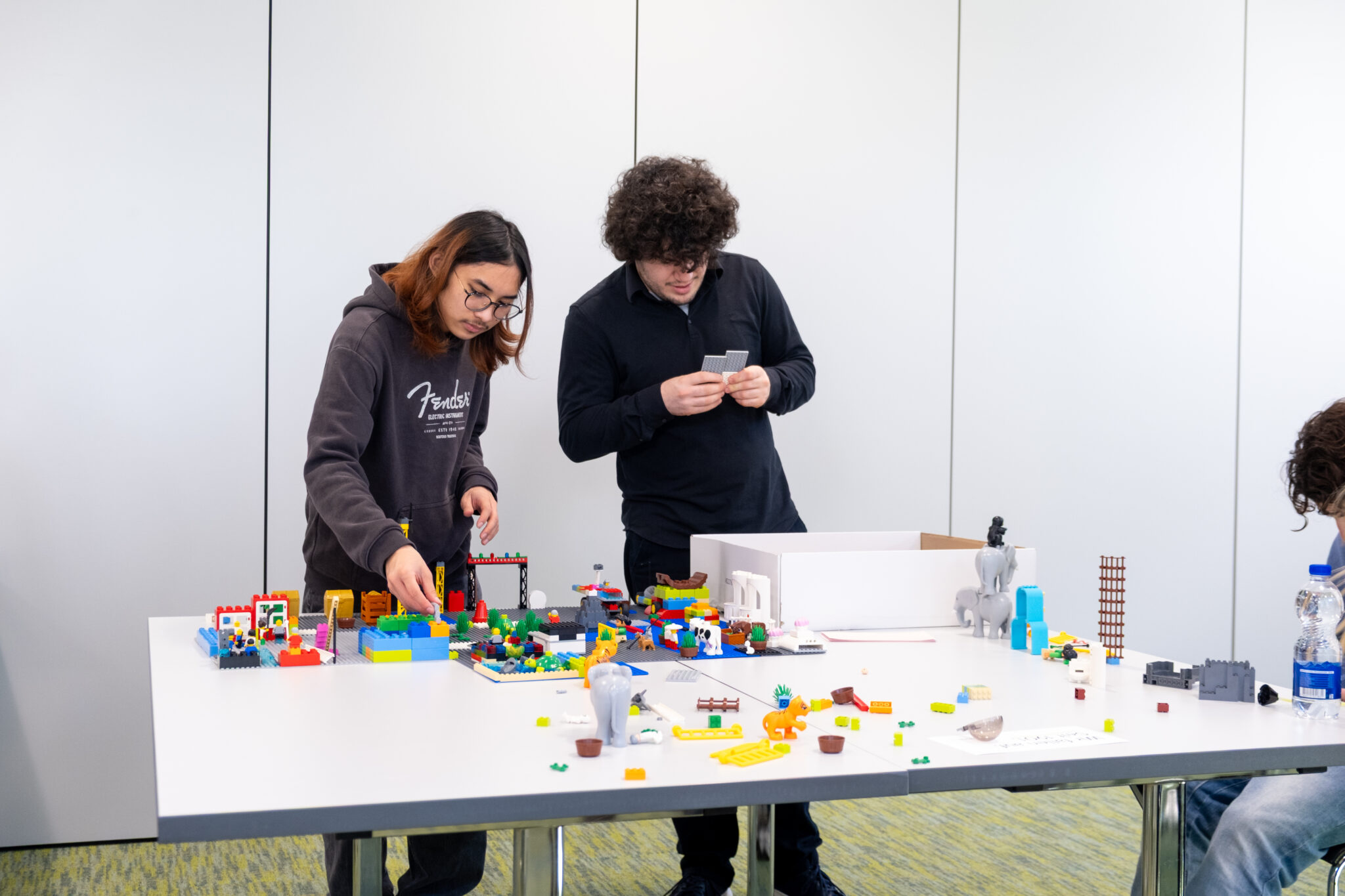 Zwei Personen stehen an einem Tisch, der mit bunten Legosteinen und -konstruktionen bedeckt ist, setzen Teile zusammen und wirken dabei ganz bei der Sache. Im Hintergrund ist eine schlichte weiße Wand zu sehen, und die Szene scheint eine kreative oder gemeinschaftliche Aktivität darzustellen.