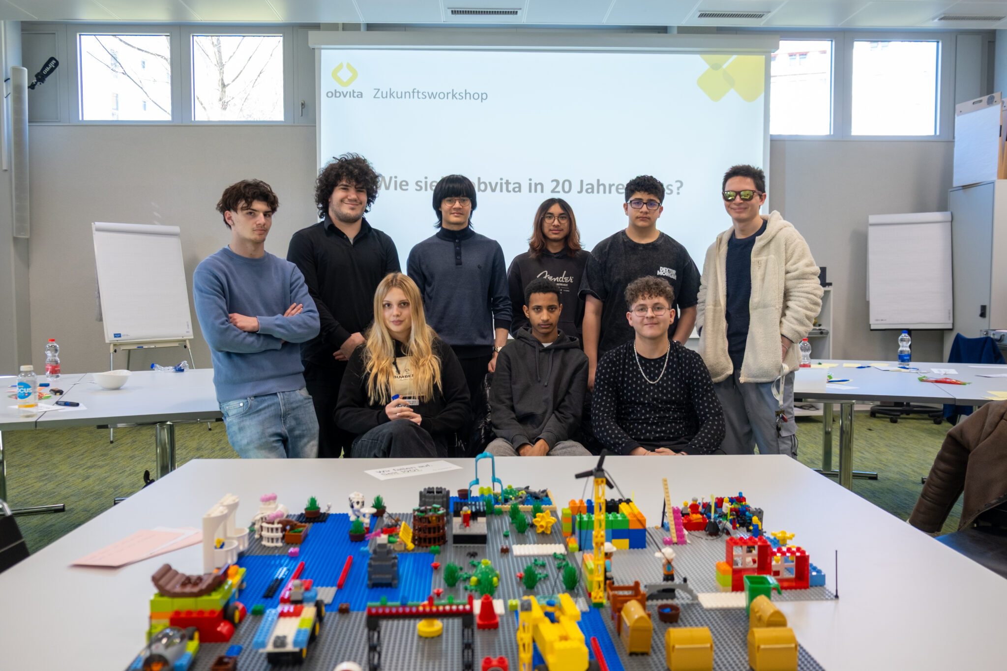 Eine Gruppe von zehn Jugendlichen posiert gemeinsam in einem hellen Raum; sie stehen und sitzen hinter einem Tisch, der mit bunten LEGO-Konstruktionen bedeckt ist. Auf einer Leinwand im Hintergrund ist ein deutscher Text zu sehen.