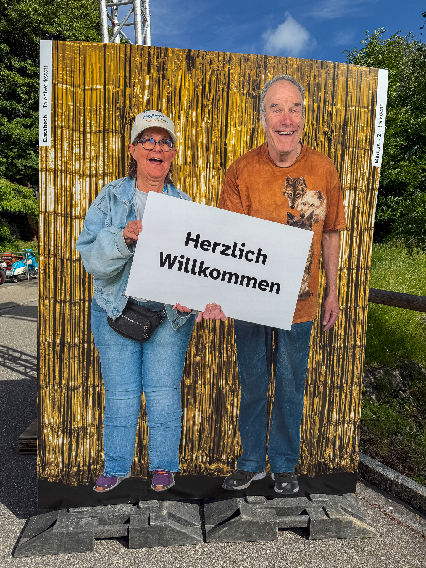 Zwei Personen stehen hinter einer Fototafel mit Ausschnitten für ihre Gesichter und halten vor einem goldenen Lametta-Hintergrund im Freien ein Schild mit der Aufschrift „Herzlich Willkommen“ (deutsch für „Warm Welcome“) in der Hand.