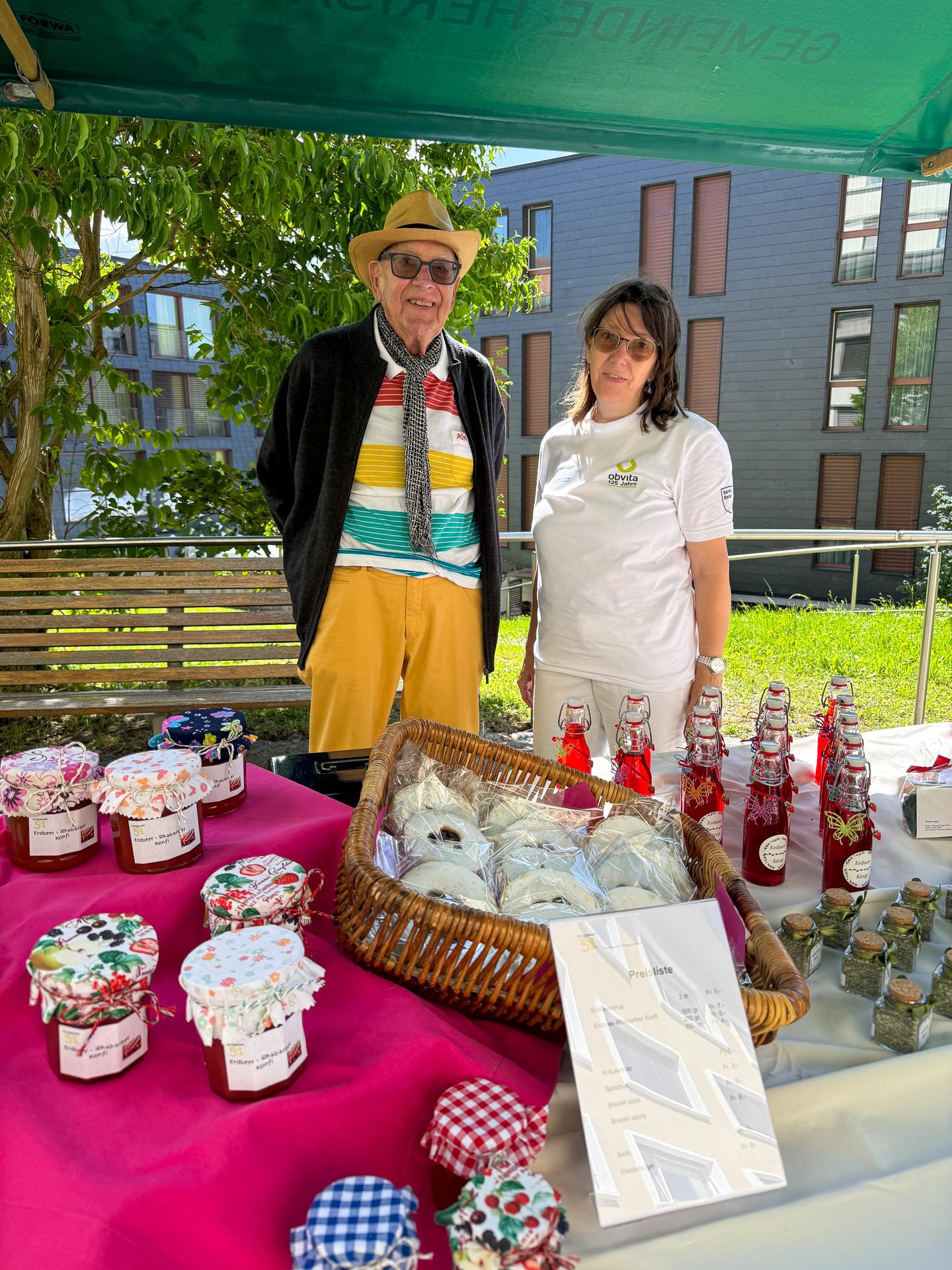 Ein älterer Mann mit Hut und buntem Hemd steht neben einer Frau in einem weißen T-Shirt hinter einem Tisch, auf dem Gläser mit Marmelade, Flaschen und Backwaren an einem Marktstand im Freien ausgestellt sind. Im Hintergrund sind Gebäude und Grünflächen zu sehen.