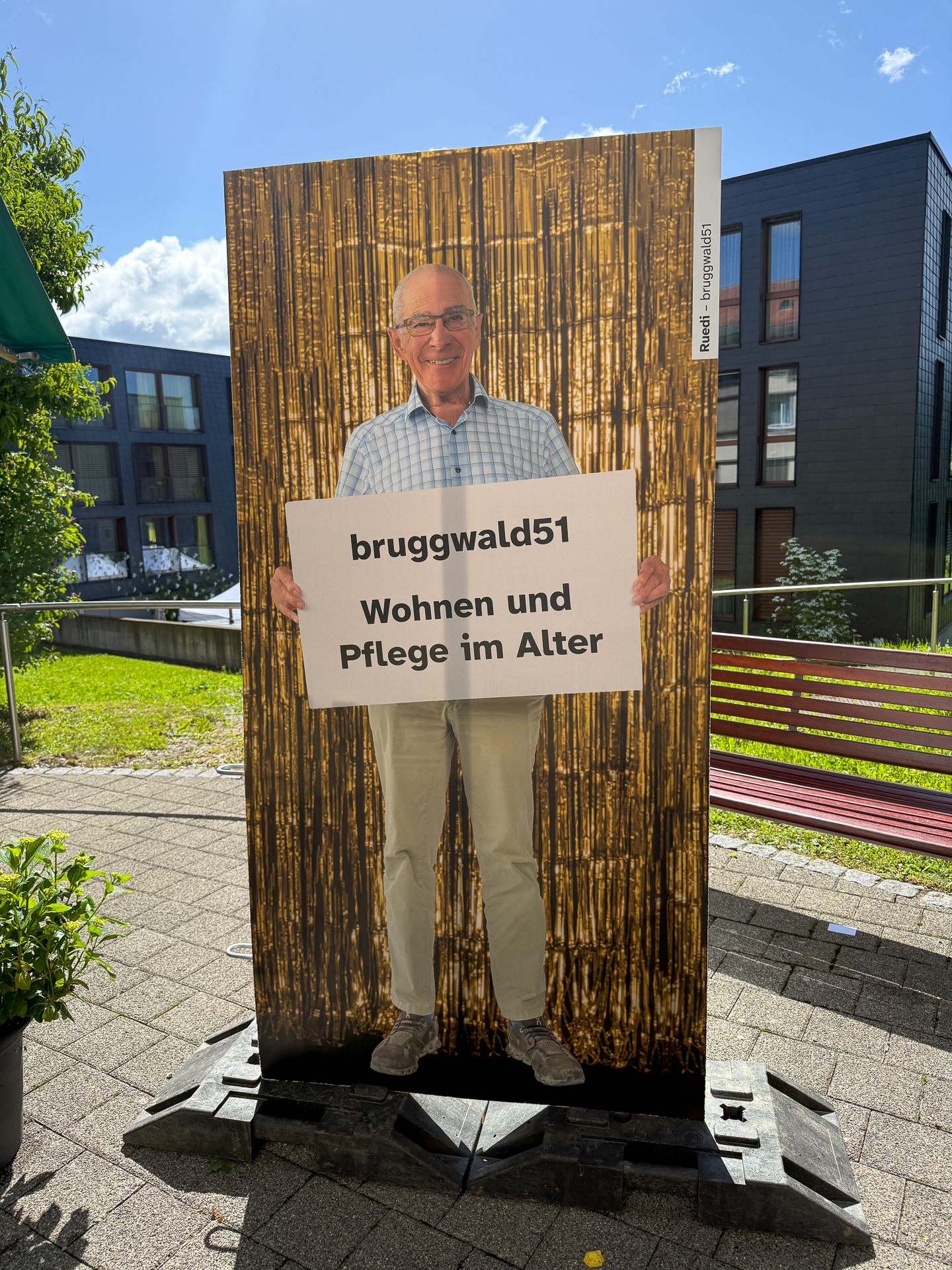 Ein Mann steht im Freien vor einem goldenen Lametta-Hintergrund und hält ein Schild mit der Aufschrift „bruggwald51 Wohnen und Pflege im Alter“ auf Deutsch in der Hand. Im Hintergrund sind moderne Gebäude und Grünflächen zu sehen.