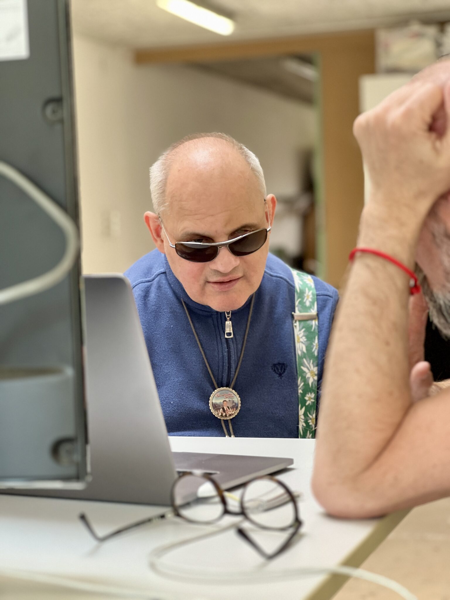 Ein Mann mit dunkler Sonnenbrille und blauem Hemd sitzt an einem Tisch und schaut auf einen Laptop. Ihm gegenüber sitzt eine weitere Person, die nur teilweise zu sehen ist; sie trägt eine Brille, und auf dem Tisch liegt ein rotes Armband.