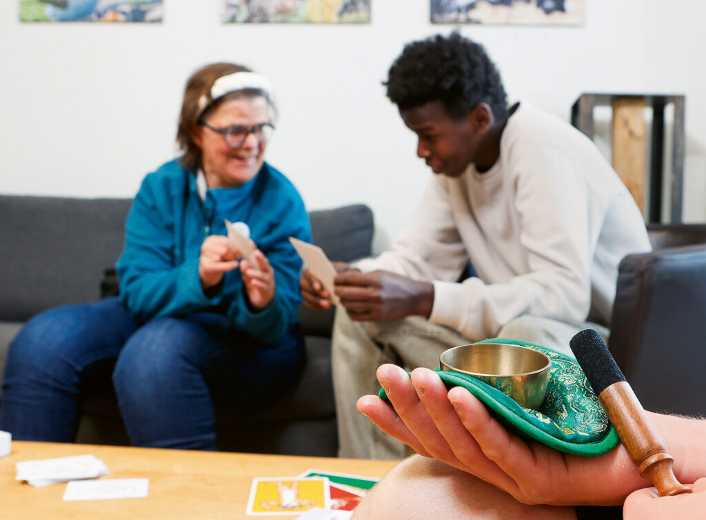 Zwei Personen sitzen auf einer Couch und schauen sich lächelnd und beschäftigt Karten an. Im Vordergrund hält eine Hand eine kleine Glocke und einen gepolsterten Schläger auf einem grünen Tuch, leicht unscharf.