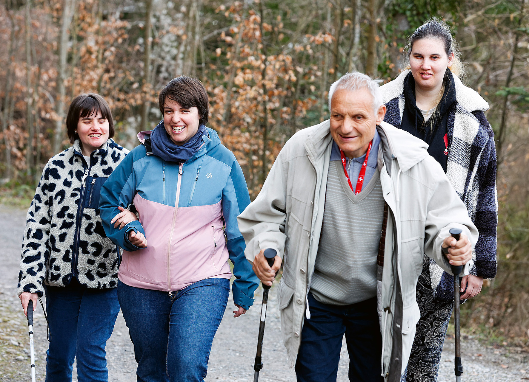 Vier Personen, drei Frauen und ein älterer Mann mit Wanderstöcken, gehen gemeinsam auf einem Waldweg, lächeln und genießen die freie Natur. Die Gruppe wirkt glücklich und ist warm gekleidet.