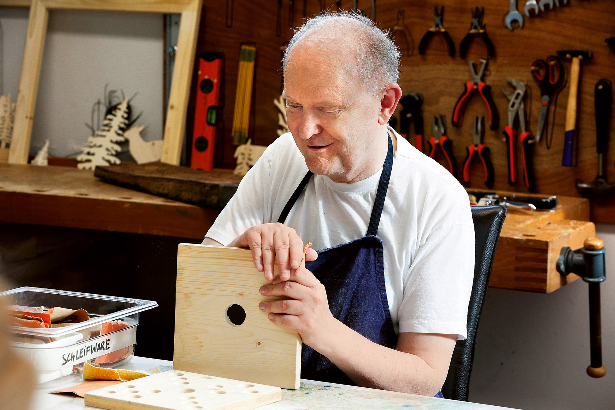 Ein Mann in einer Werkstatt, der ein weißes Hemd und eine dunkle Schürze trägt, hält ein Holzbrett mit einem Loch in der Hand. Hinter ihm hängen verschiedene Werkzeuge an der Wand, und auf dem Tisch liegen weitere Holzgegenstände.