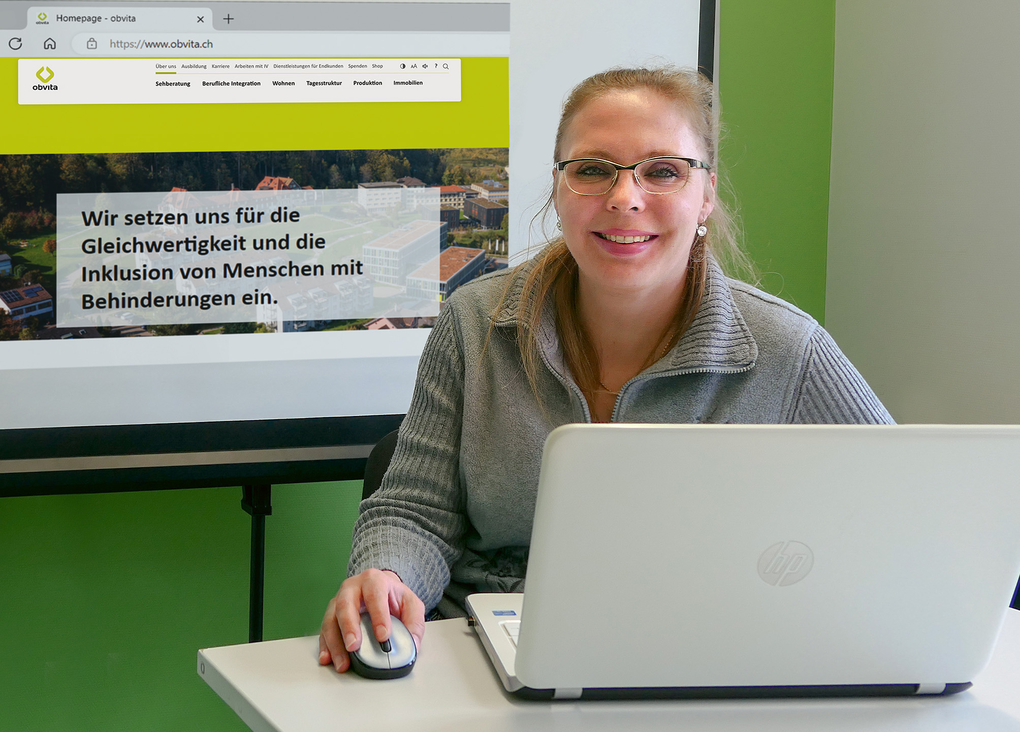 Eine Frau mit Brille und grauem Pullover sitzt an einem Schreibtisch mit Laptop und Maus und lächelt. Hinter ihr zeigt ein Bildschirm eine Website, die für Inklusion und Gleichstellung von Menschen mit Behinderungen wirbt.