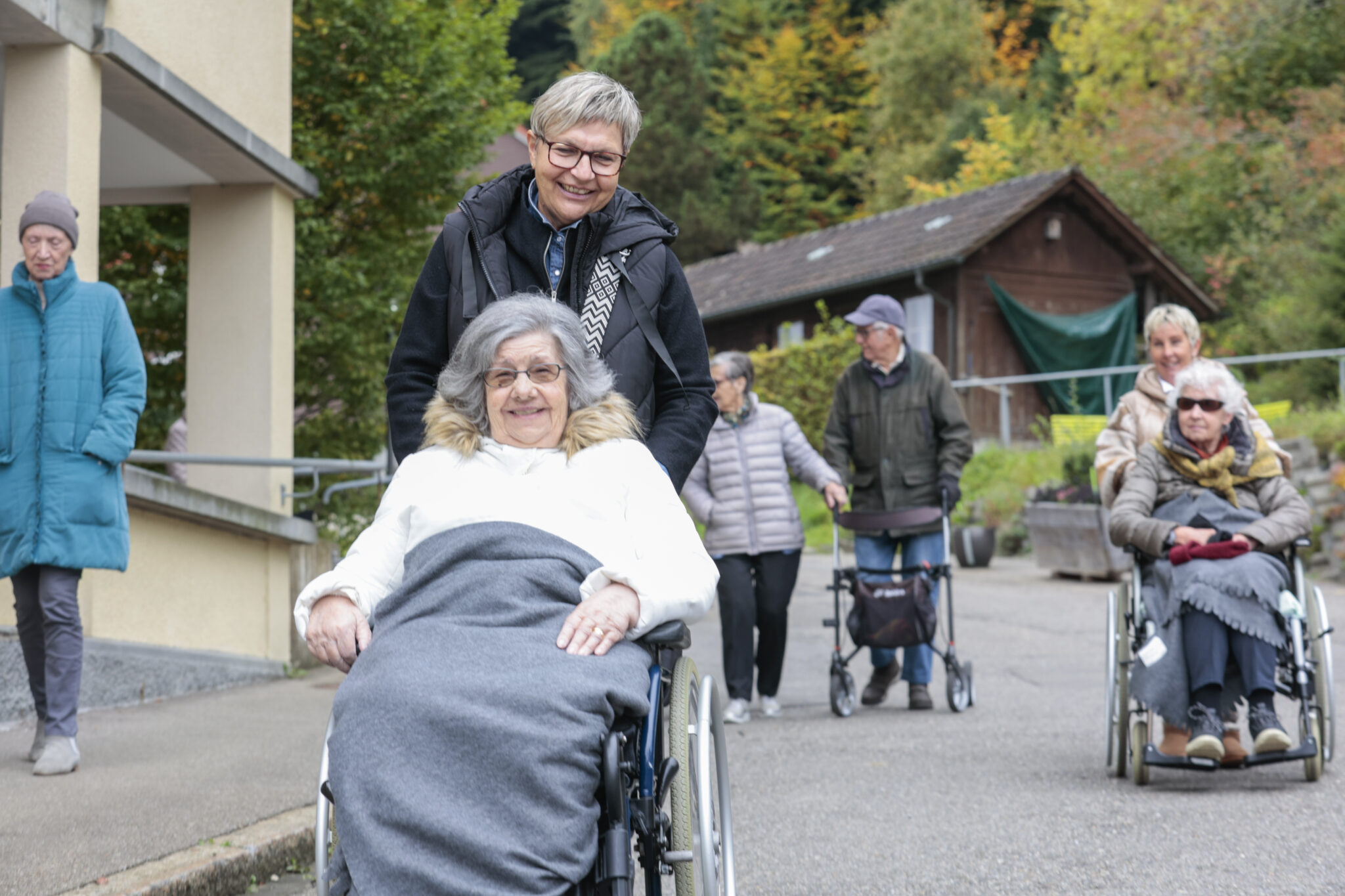 Eine lächelnde Frau schiebt eine ältere Frau im Rollstuhl ins Freie, während andere Senioren in der Nähe spazieren gehen. Im Hintergrund sind Bäume und ein kleines Gebäude zu sehen.