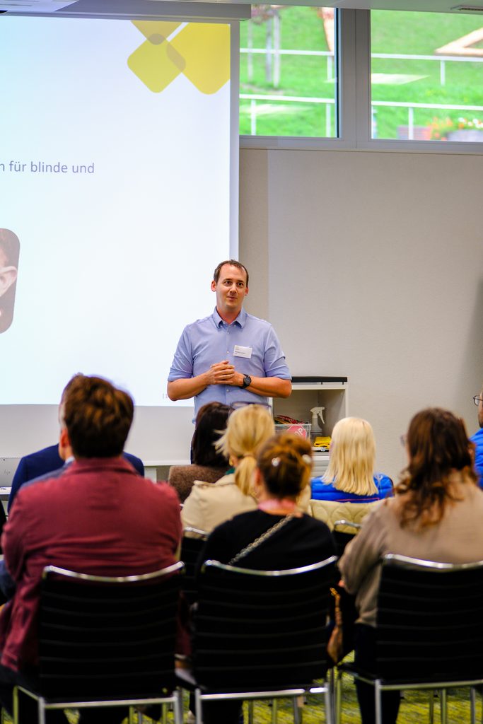Ein Mann steht vor einem Publikum, das auf Stühlen sitzt, und hält eine Rede; im Hintergrund sind eine Präsentationsleinwand und ein Fenster zu sehen.