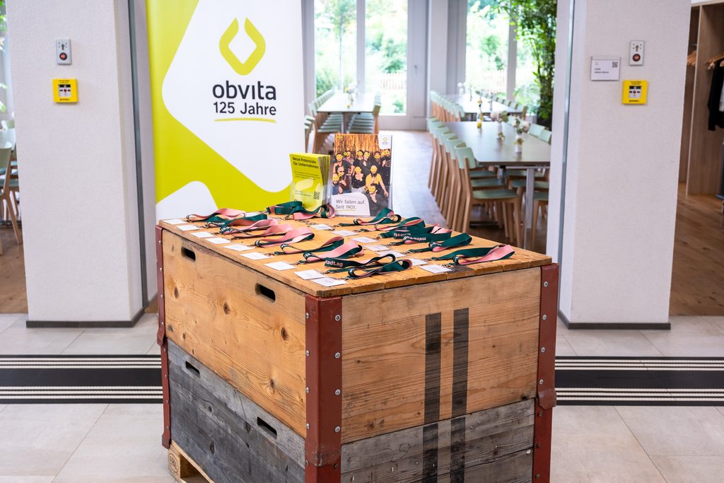 Ein Holztisch mit Medaillen, die mit grünen und rosa Bändern verziert sind, ein Schild mit der Aufschrift „obvita 125 Jahre“ und eine Fotoausstellung in einem hellen Raum mit langen Tischen und Stühlen.