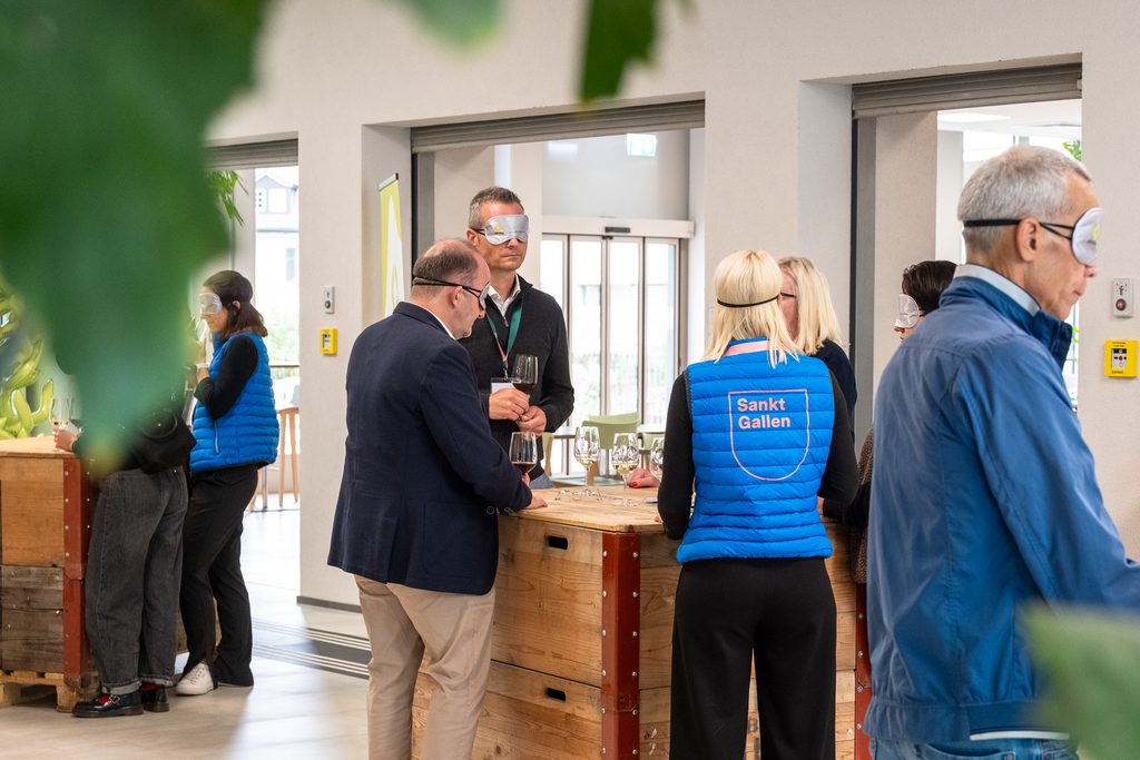 Menschen mit Augenbinden und blauen „Sankt Gallen“-Westen stehen drinnen um Holztische herum und nehmen an einer Weinverkostung oder einer sensorischen Aktivität teil. Einige von ihnen halten Gläser in der Hand, und im Vordergrund sind grüne Pflanzen zu sehen.