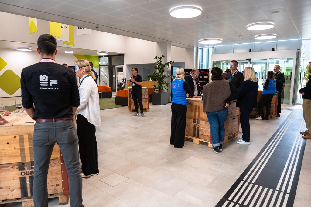 In einem modernen Innenraum mit Holztischen, heller Beleuchtung und Schildern an den Wänden stehen Menschen und unterhalten sich. Einige tragen Namensschilder und Westen mit Logos, was auf eine Networking-Veranstaltung oder ein gesellschaftliches Event hindeutet.