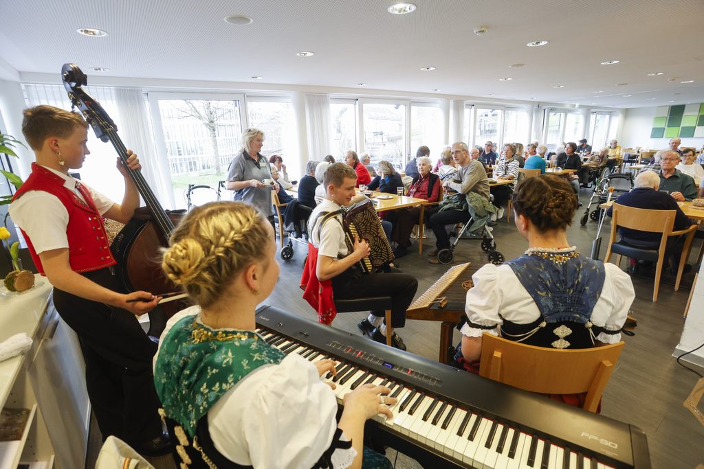 Eine Gruppe junger Musiker in traditioneller Tracht spielt vor einem sitzenden Publikum in einem hellen, modernen Raum, während die Zuschauer die Musik verfolgen und genießen.