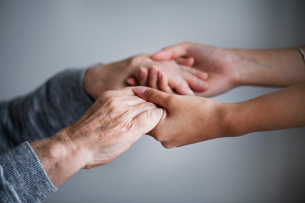 Vor einem schlichten, sanft beleuchteten Hintergrund hält eine jüngere Person behutsam die Hände einer älteren Person in den Händen und vermittelt so Fürsorge und Unterstützung.