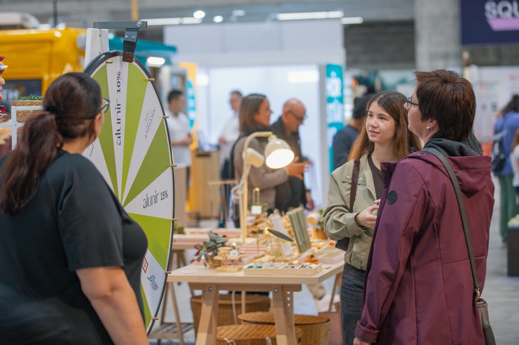 Eine Person an einem Messestand unterhält sich mit zwei Besuchern neben einem sich drehenden Glücksrad mit der Aufschrift „alunir“.
