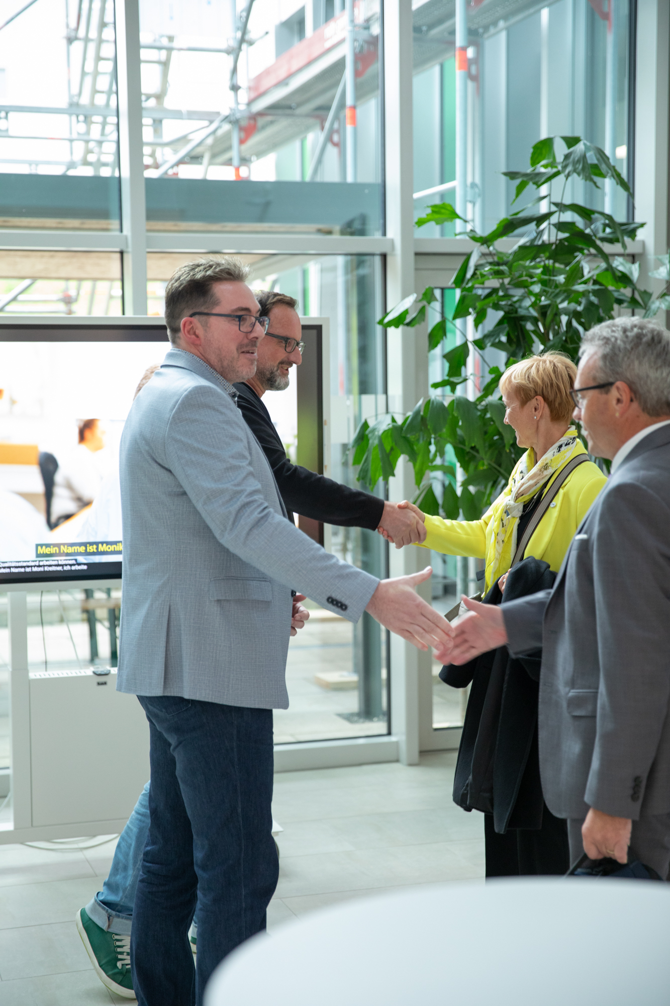 Zwei Männer in Anzügen geben sich die Hand, während eine Frau in einer gelben Jacke eine andere Person in der Lobby eines Büros begrüßt.