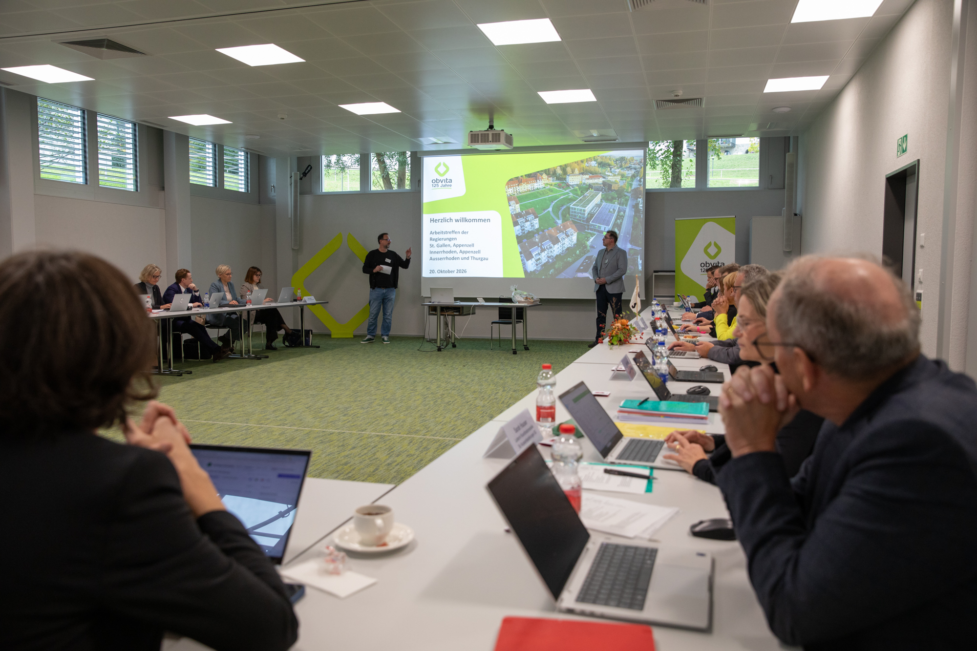 Zwei Redner stehen vorne im Konferenzraum und halten einen Vortrag vor einer Gruppe, die an langen Tischen sitzt, während auf einer Leinwand „Herzlich willkommen“ sowie die Details zur Veranstaltung zu sehen sind.