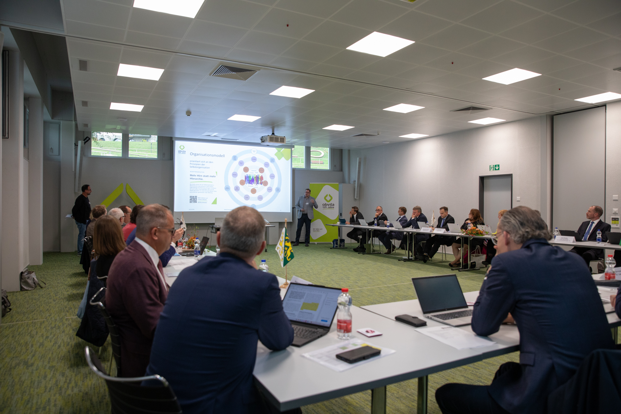 Ein Redner präsentiert einer Gruppe von Personen, die an langen Tischen in einem Konferenzraum sitzen, ein Organisationsmodell auf einer Leinwand.