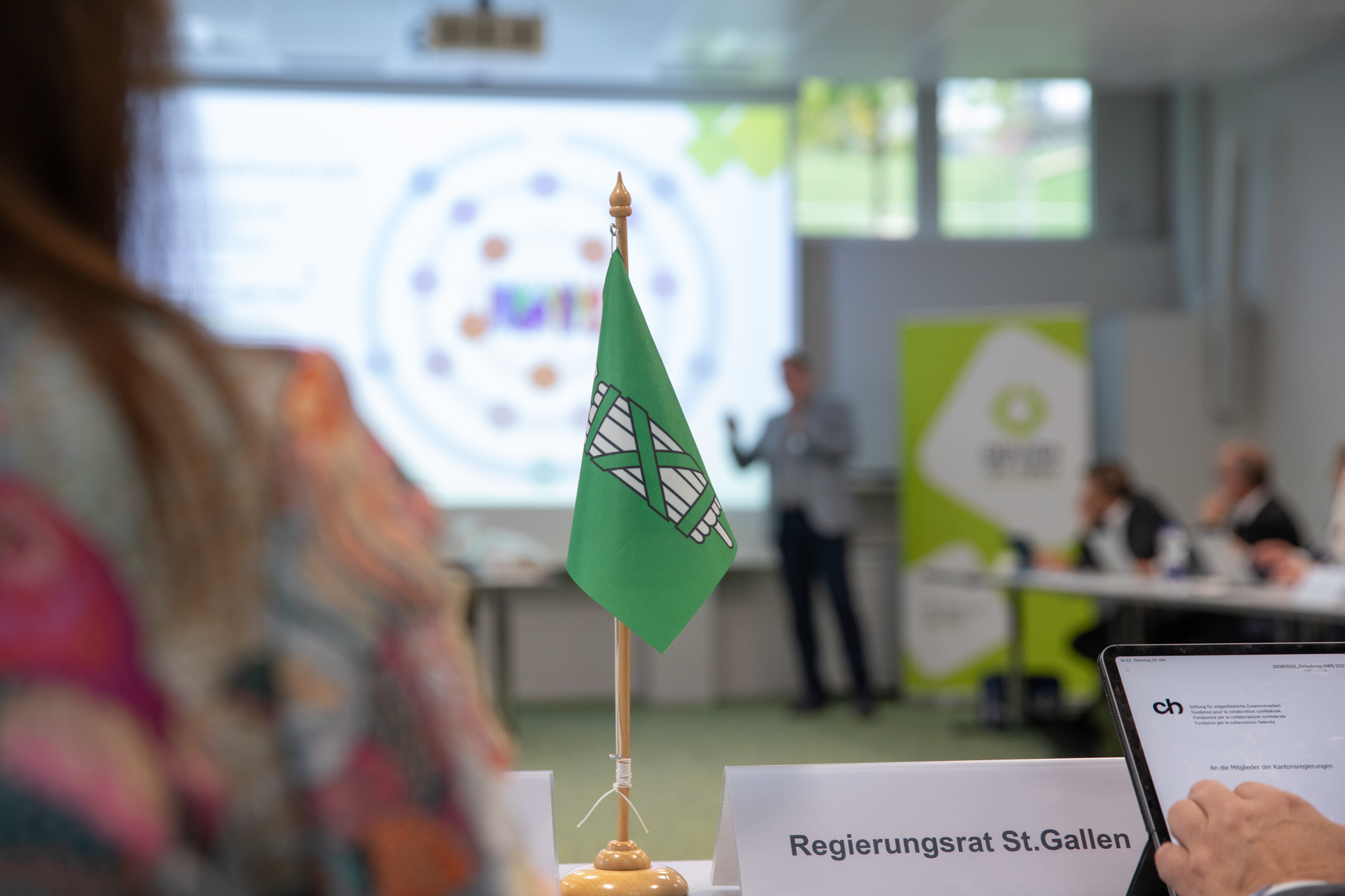 Auf einem Konferenztisch in einem Besprechungsraum steht eine kleine grüne Flagge mit einem weißen Wappen vor einem Namensschild mit der Aufschrift „Regierungsrat St. Gallen“.