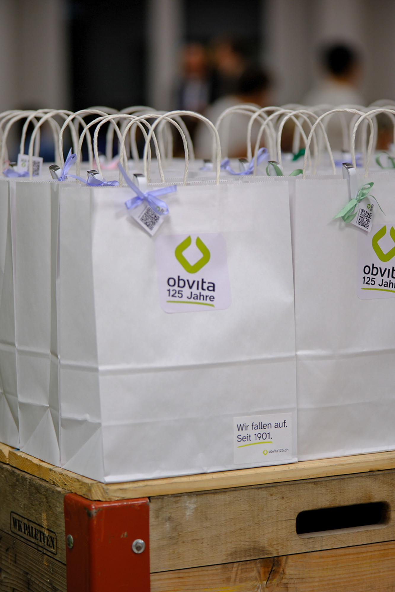 Reihen von weißen Geschenktüten aus Papier mit dem „obvita 125 Jahre“-Logo, gestapelt auf einer Holzpalette.