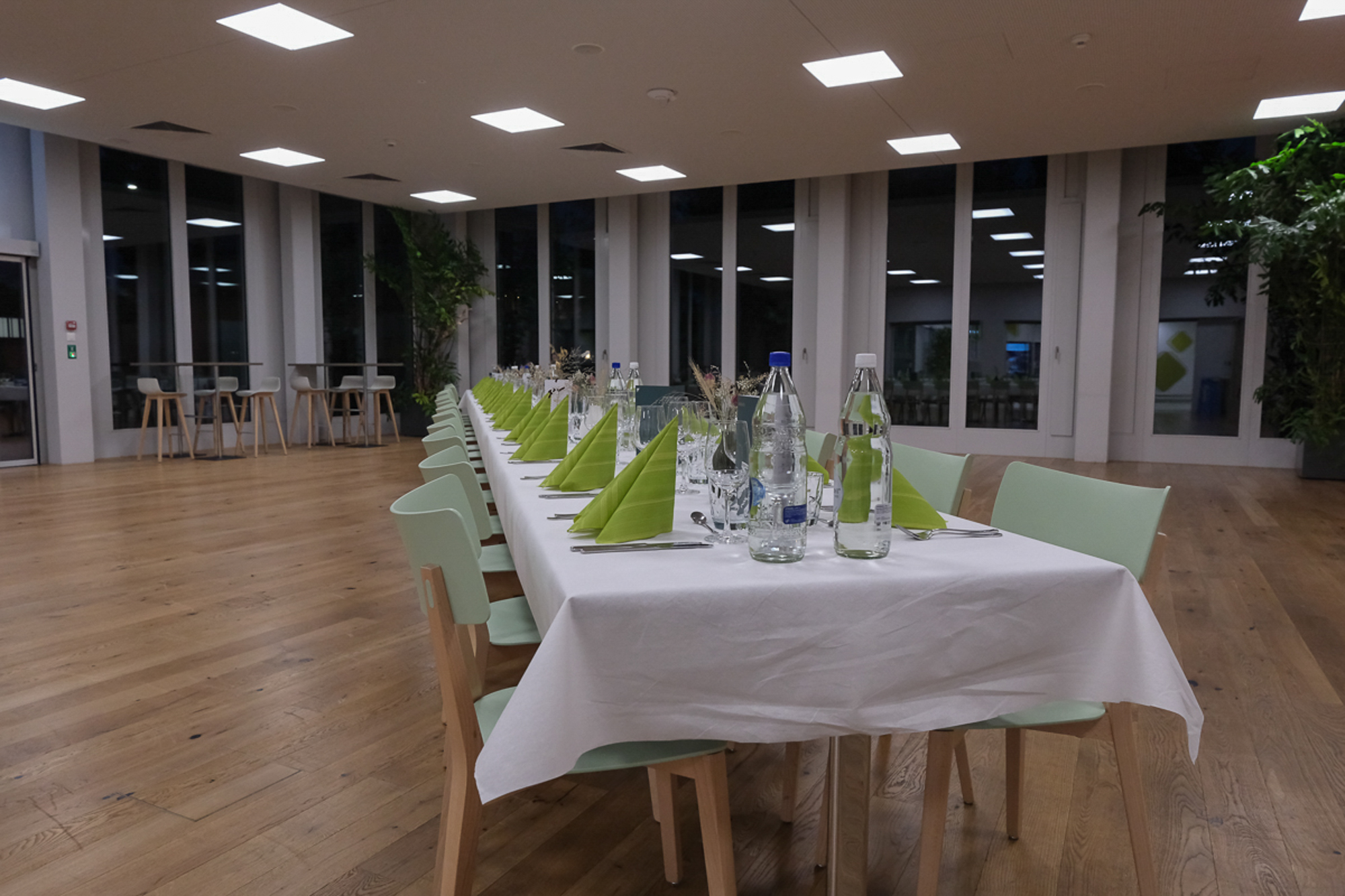 Ein langer Esstisch mit weißen Tischdecken, hellgrünen Servietten, Gläsern und Wasserflaschen in einem geräumigen Saal.