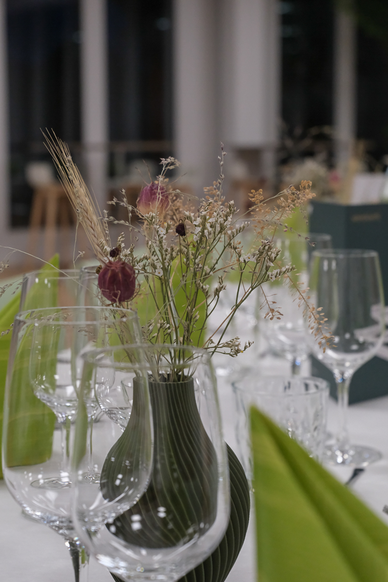 Ein Tischschmuck aus getrockneten Gräsern und Blumen in einer grünen, geriffelten Vase auf einem Tisch, der mit Weingläsern und gefalteten grünen Servietten gedeckt ist.