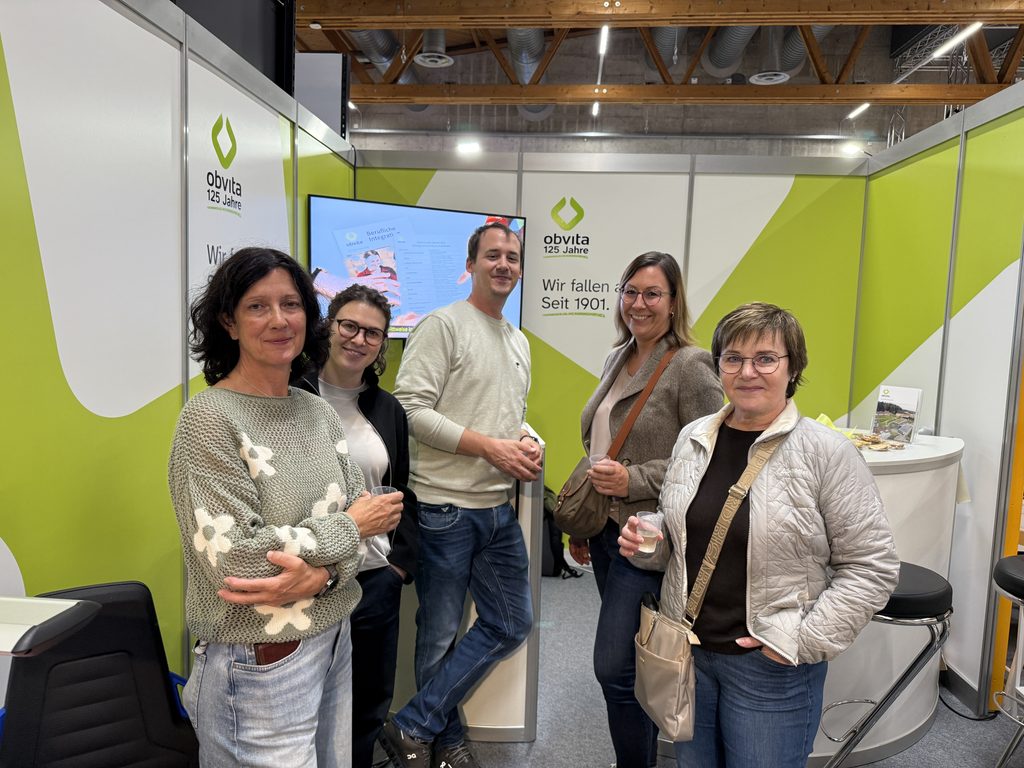 Fünf Erwachsene posieren gemeinsam an einem Messestand, der mit dem Logo „Obvita 125 Jahre“ versehen ist.